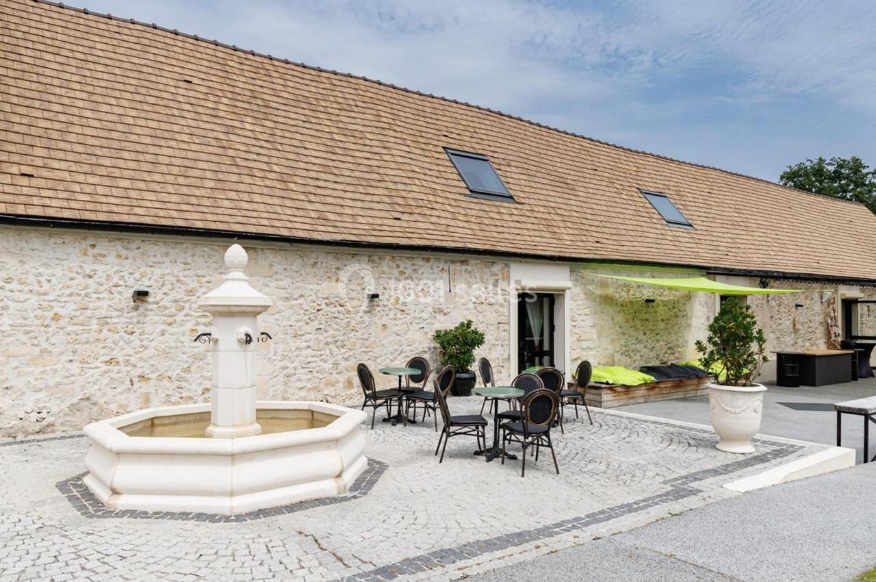 Cour pavée avec une fontaine centrale, entourée de tables et chaises, devant un bâtiment en pierre avec toit en tuiles.