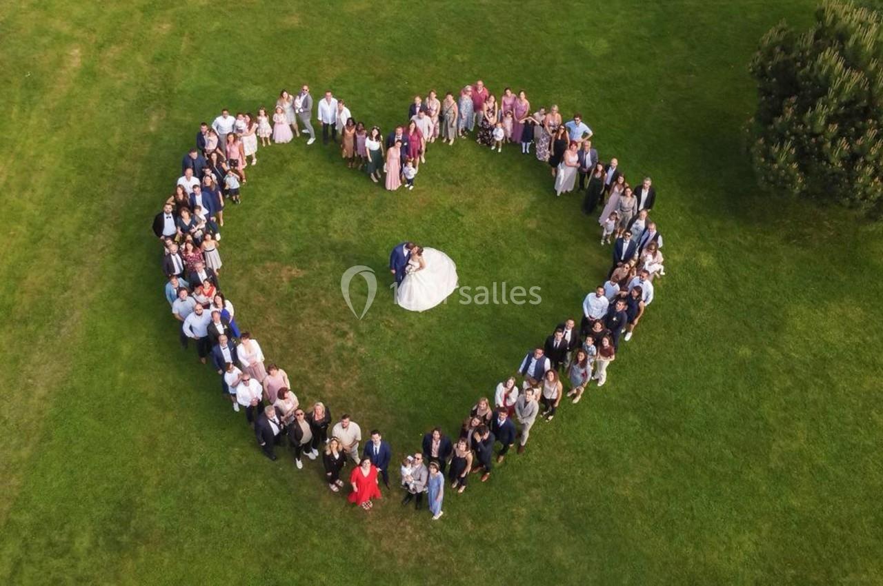 Groupe de personnes formant un cœur autour d'un couple en tenue de mariage sur une pelouse verte.