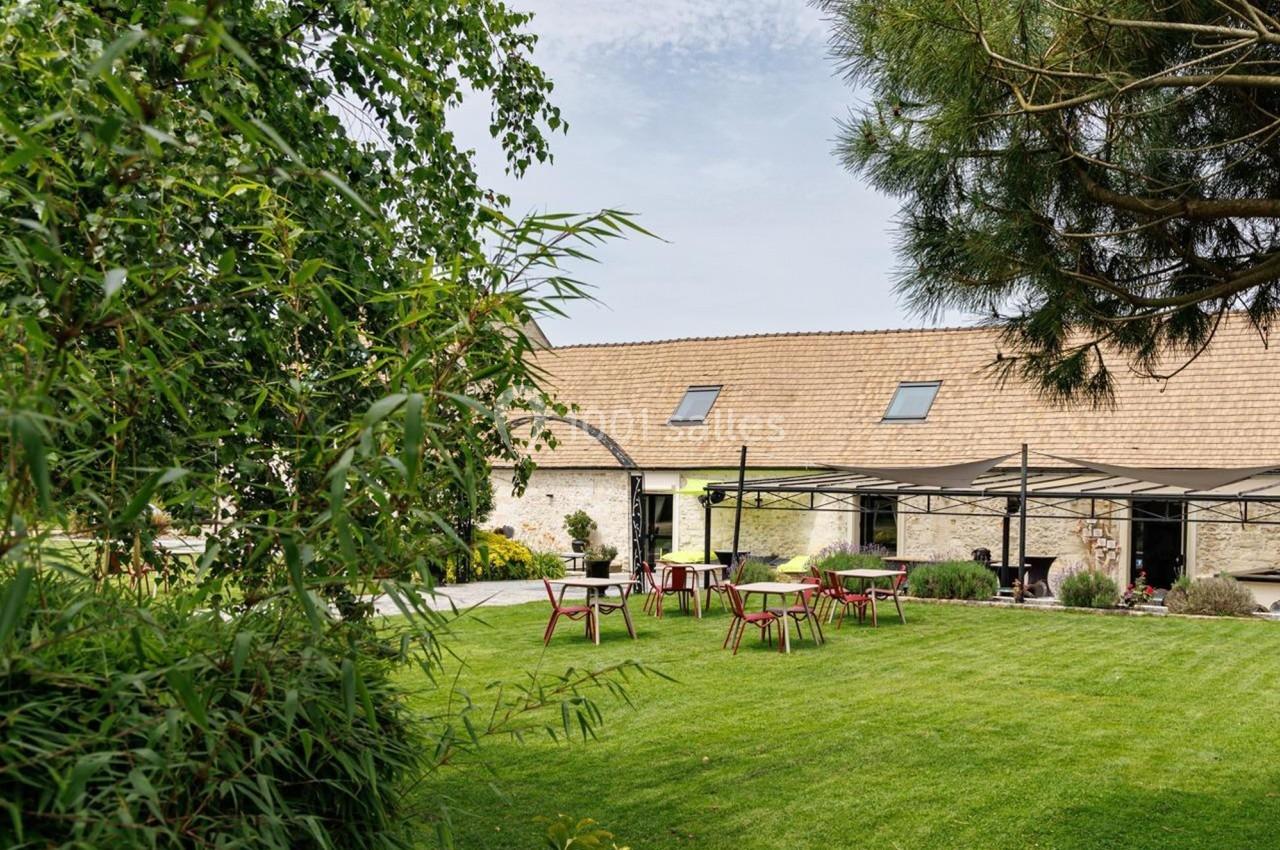 Jardin verdoyant avec pelouse, tables et chaises en bois, entouré de végétation et d'un bâtiment en pierre.