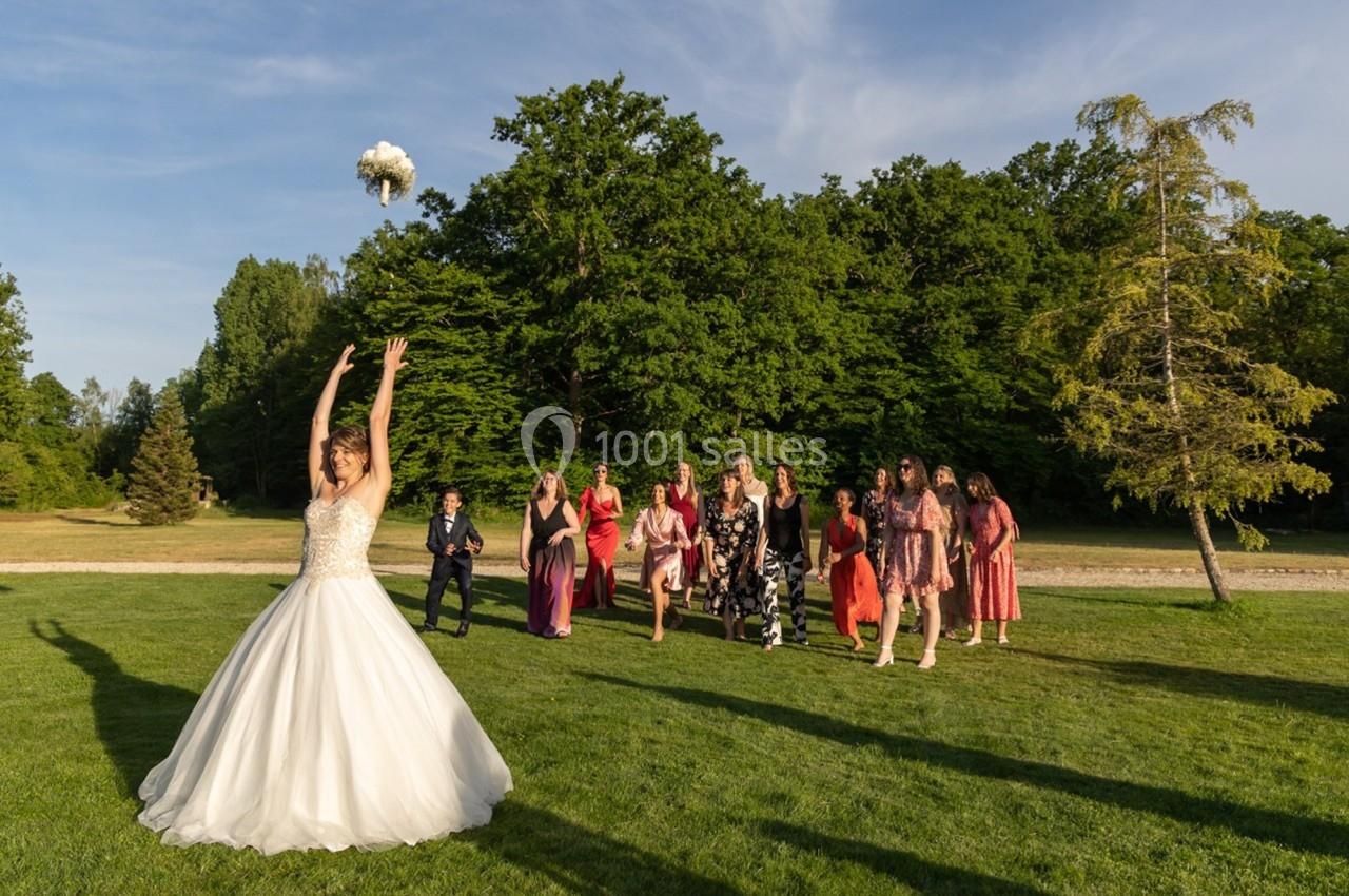 Une mariée lance son bouquet en l'air devant un groupe d'invités prêts à l'attraper, dans un parc verdoyant.