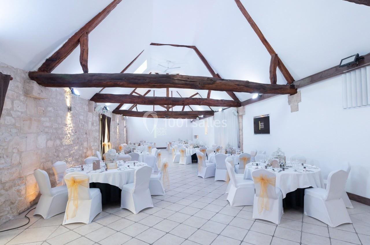 Salle de réception lumineuse avec poutres apparentes, tables rondes dressées et chaises blanches décorées de nœuds.