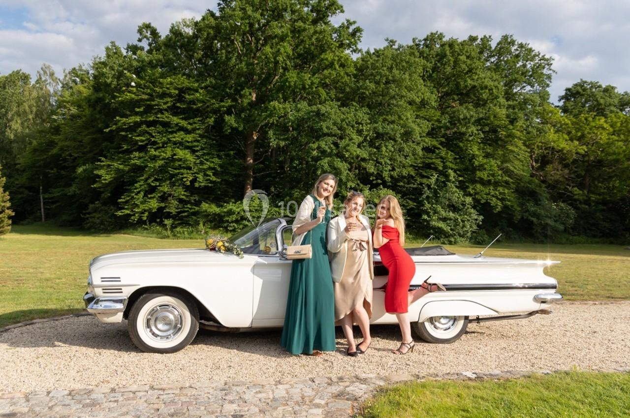 Trois femmes posent souriantes devant une voiture ancienne blanche décapotable, dans un parc verdoyant.