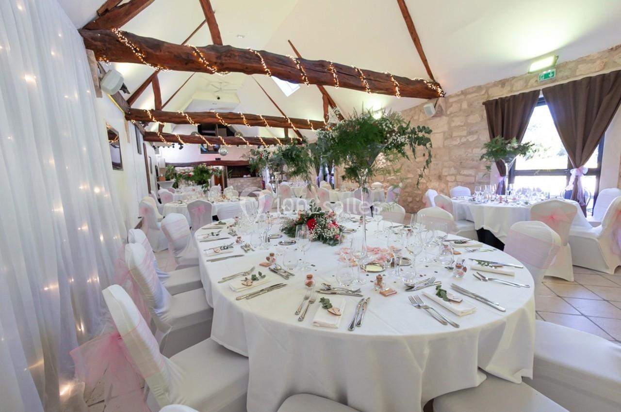Salle de réception décorée avec des tables rondes dressées, chaises blanches et guirlandes lumineuses au plafond.