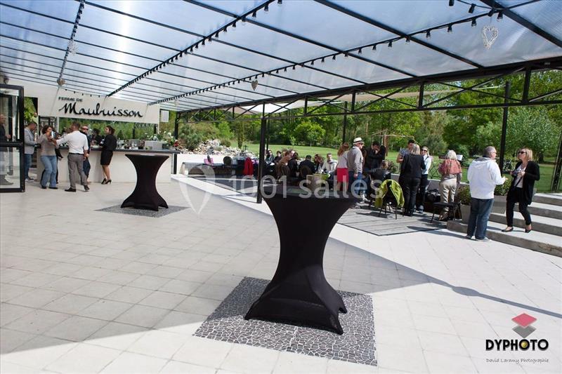 Terrasse couverte avec des tables hautes noires, des groupes de personnes discutent près d'un espace vert en arrière-plan.