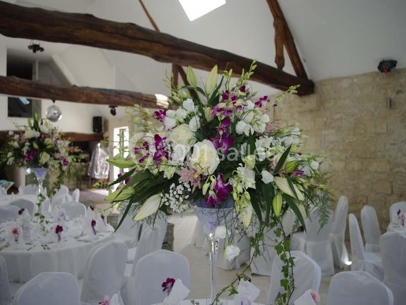 Centre de table floral avec des lys blancs, des roses et des orchidées violettes dans une salle de réception lumineuse.