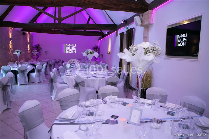 Salle de réception décorée avec des tables rondes dressées, éclairage violet et poutres apparentes au plafond.