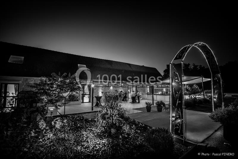 Vue nocturne en noir et blanc d'une terrasse éclairée, entourée de végétation et d'une arche lumineuse.