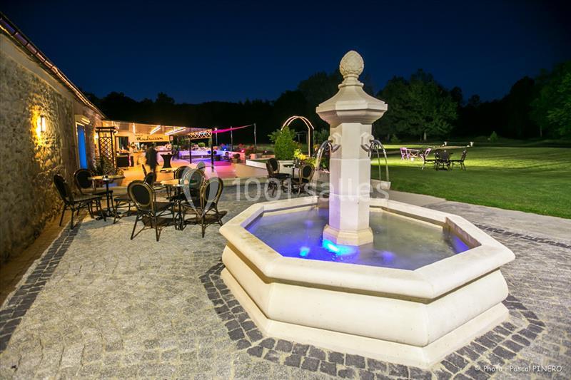 Fontaine illuminée entourée de tables et chaises sur une terrasse pavée, avec un jardin en arrière-plan, de nuit.
