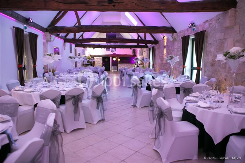 Salle de réception décorée avec des tables rondes dressées, chaises blanches et poutres apparentes sous éclairage violet.