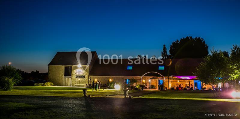 Bâtiment rural éclairé de nuit avec des lumières colorées, entouré d'un grand espace vert et d'arbres.