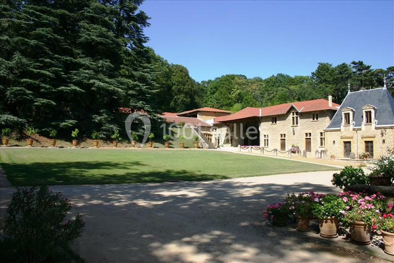 Cour d'un domaine avec pelouse, bâtiments en pierre, toits rouges, pots de fleurs et arbres en arrière-plan.