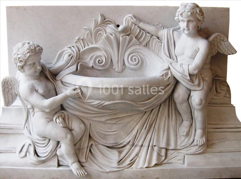 Deux angelots sculptés en marbre soutiennent une vasque ornée de motifs floraux et drapés.
