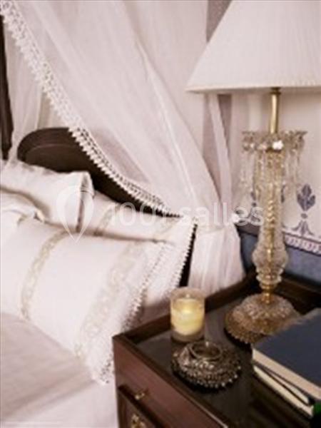 Lit avec baldaquin blanc, table de chevet en bois avec lampe en cristal, bougie et livres posés dessus.