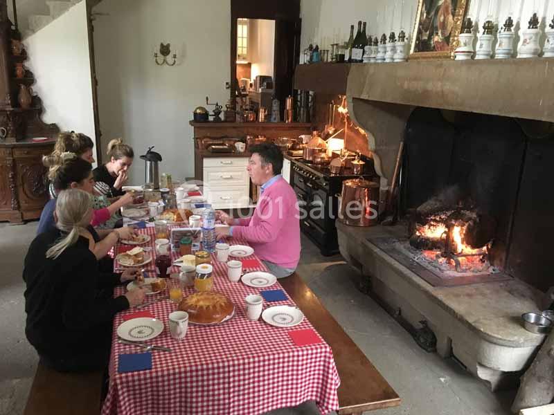 Des personnes partagent un repas autour d'une grande table dans une cuisine rustique avec une cheminée allumée.