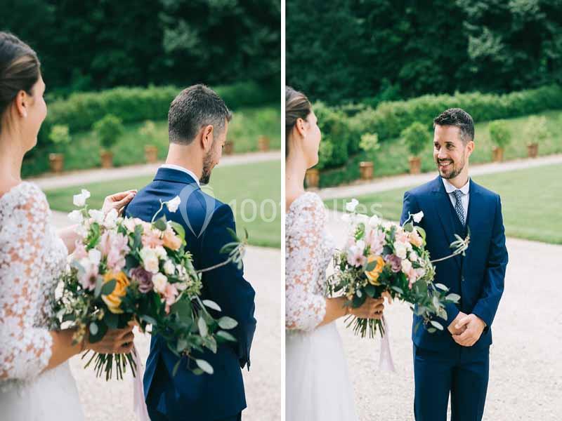 Une mariée tenant un bouquet de fleurs surprend un homme en costume bleu dans un jardin.