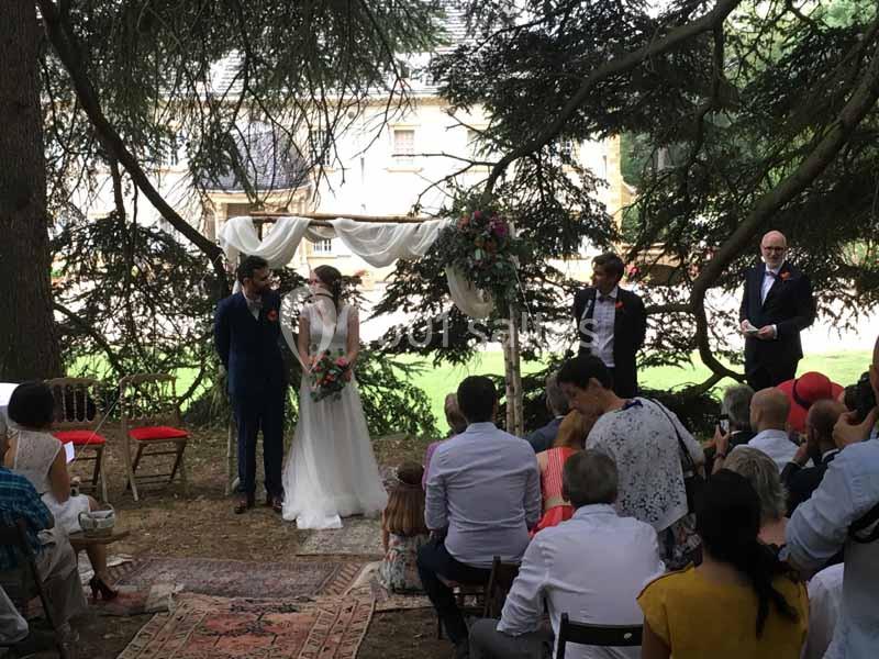 Un couple échange ses vœux de mariage en extérieur sous une arche décorée, entouré d'invités assis.