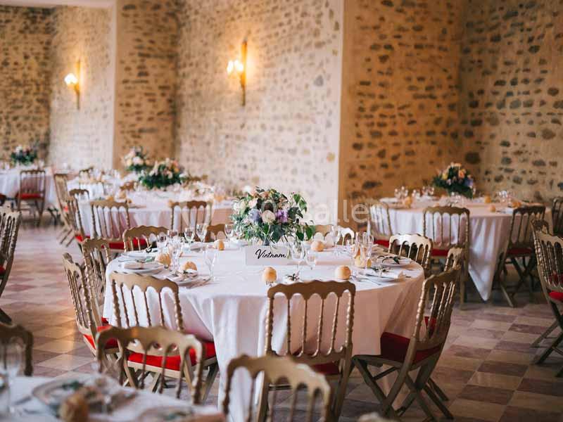 Salle de réception avec des tables rondes décorées de nappes blanches, bouquets de fleurs et chaises en bois.
