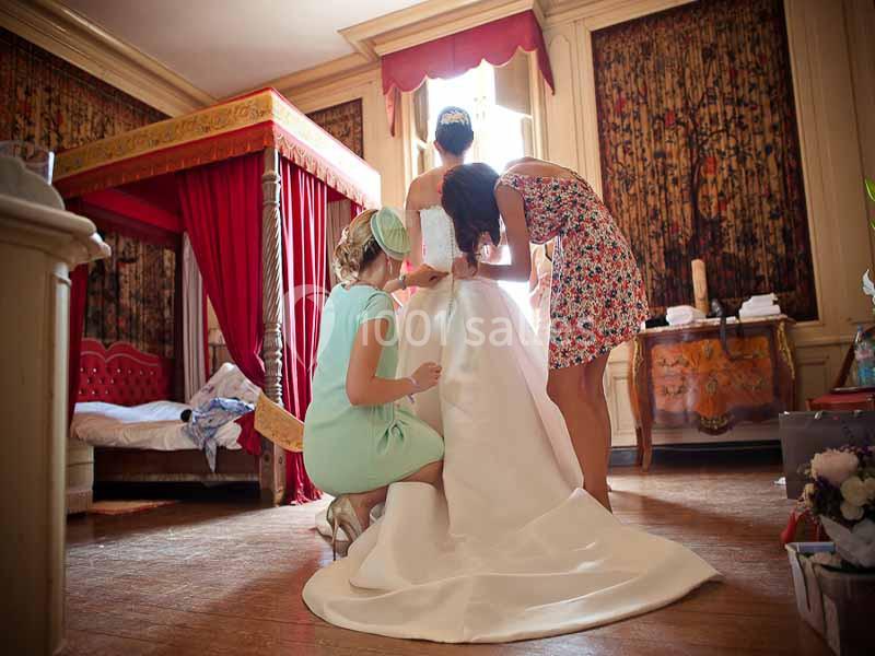 Deux femmes aident une mariée à ajuster sa robe blanche dans une chambre décorée avec un lit à baldaquin.