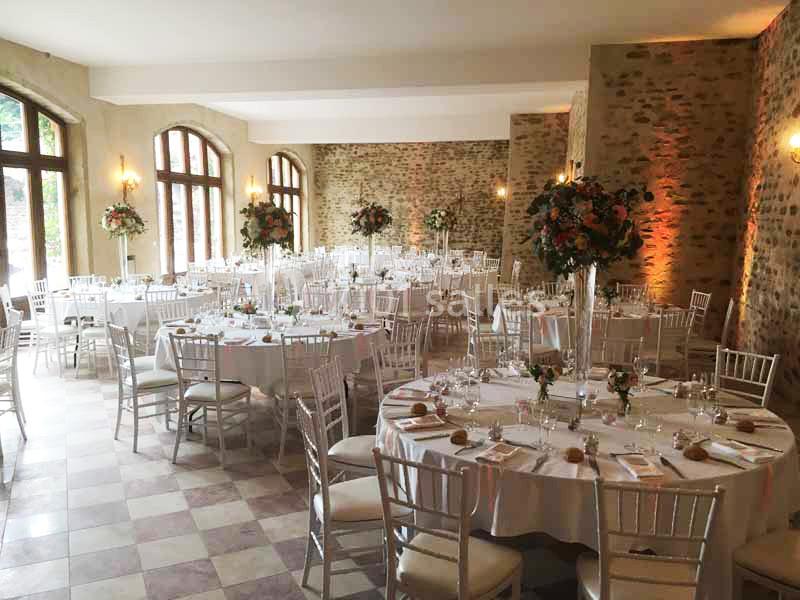 Salle de réception décorée avec des tables rondes dressées, chaises blanches et arrangements floraux élégants.