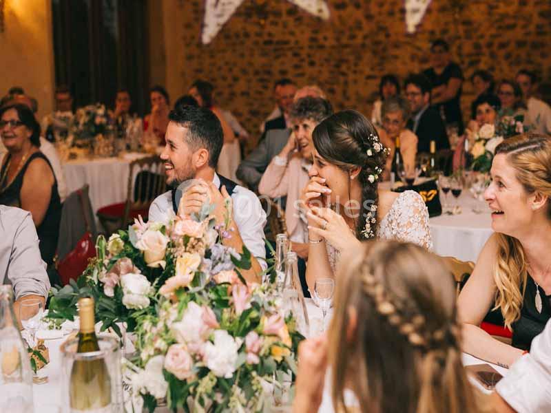 Des invités rient et discutent autour de tables décorées de fleurs lors d'une réception festive en intérieur.