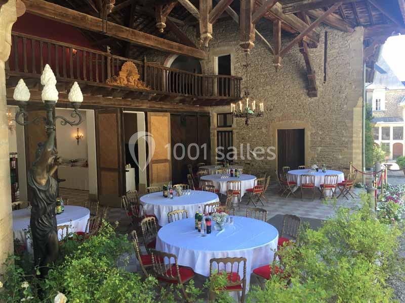 Salle extérieure avec tables rondes dressées, chaises en bois et décor rustique sous une charpente en bois.
