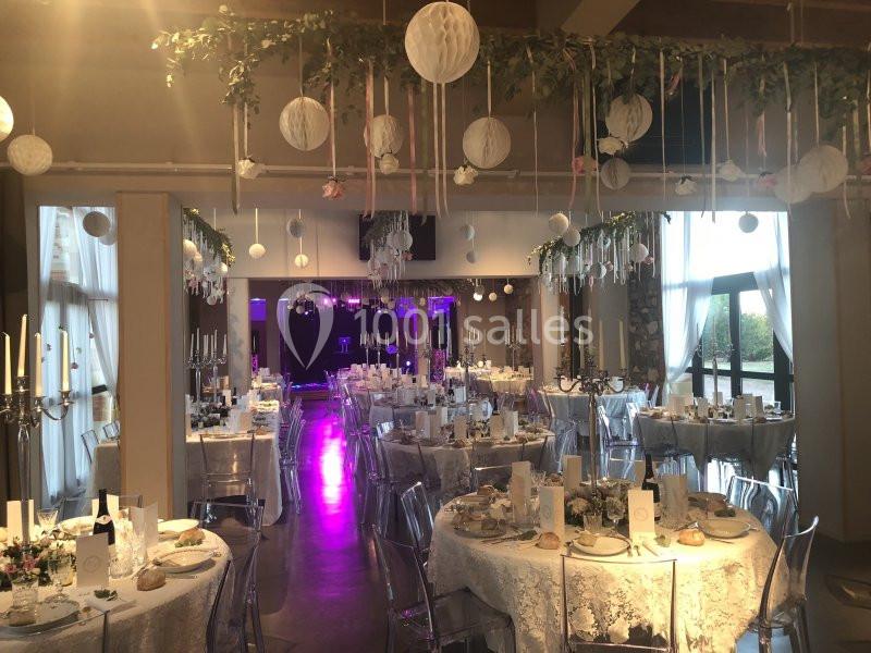 Salle de réception décorée pour un mariage, avec tables dressées, chandeliers et suspensions florales.