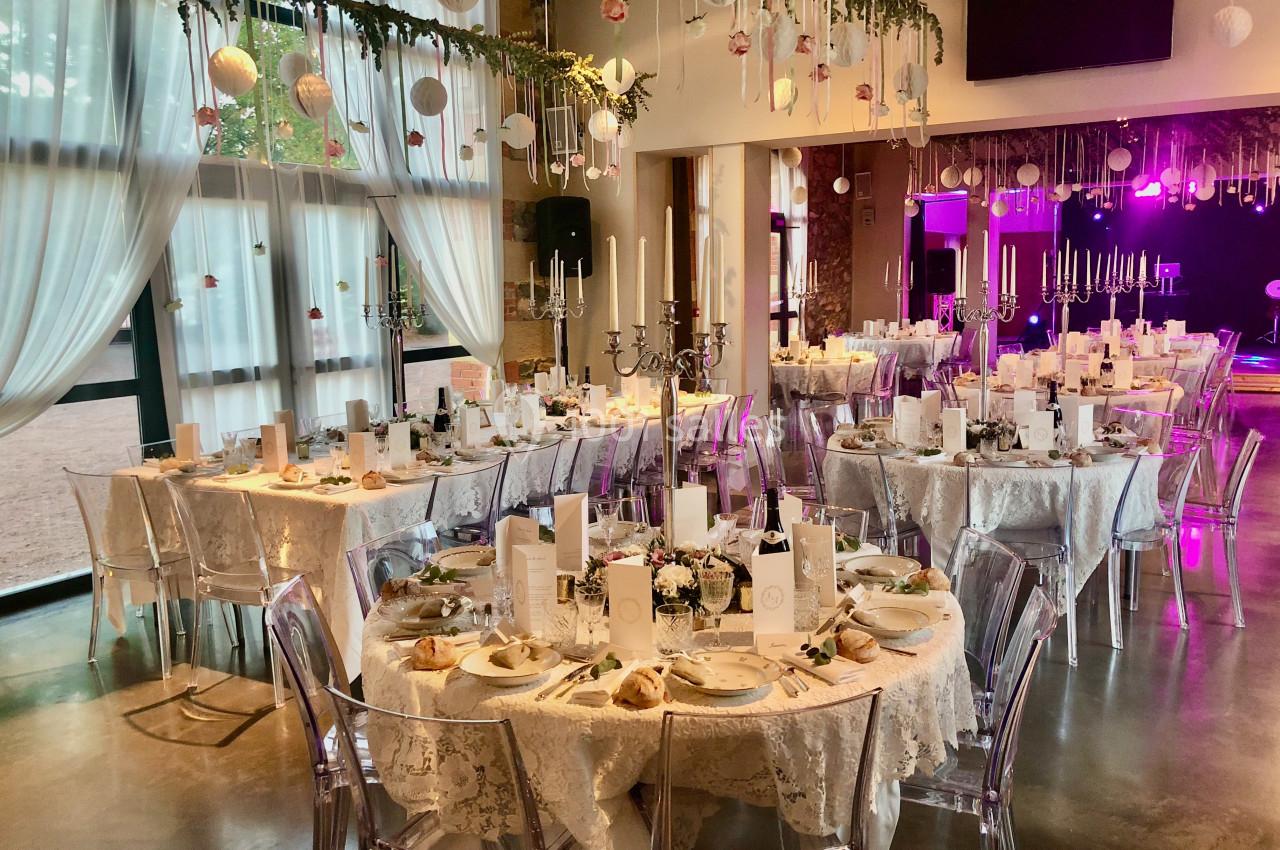 Salle de réception décorée pour un événement, avec tables rondes dressées, nappes blanches et chandeliers élégants.