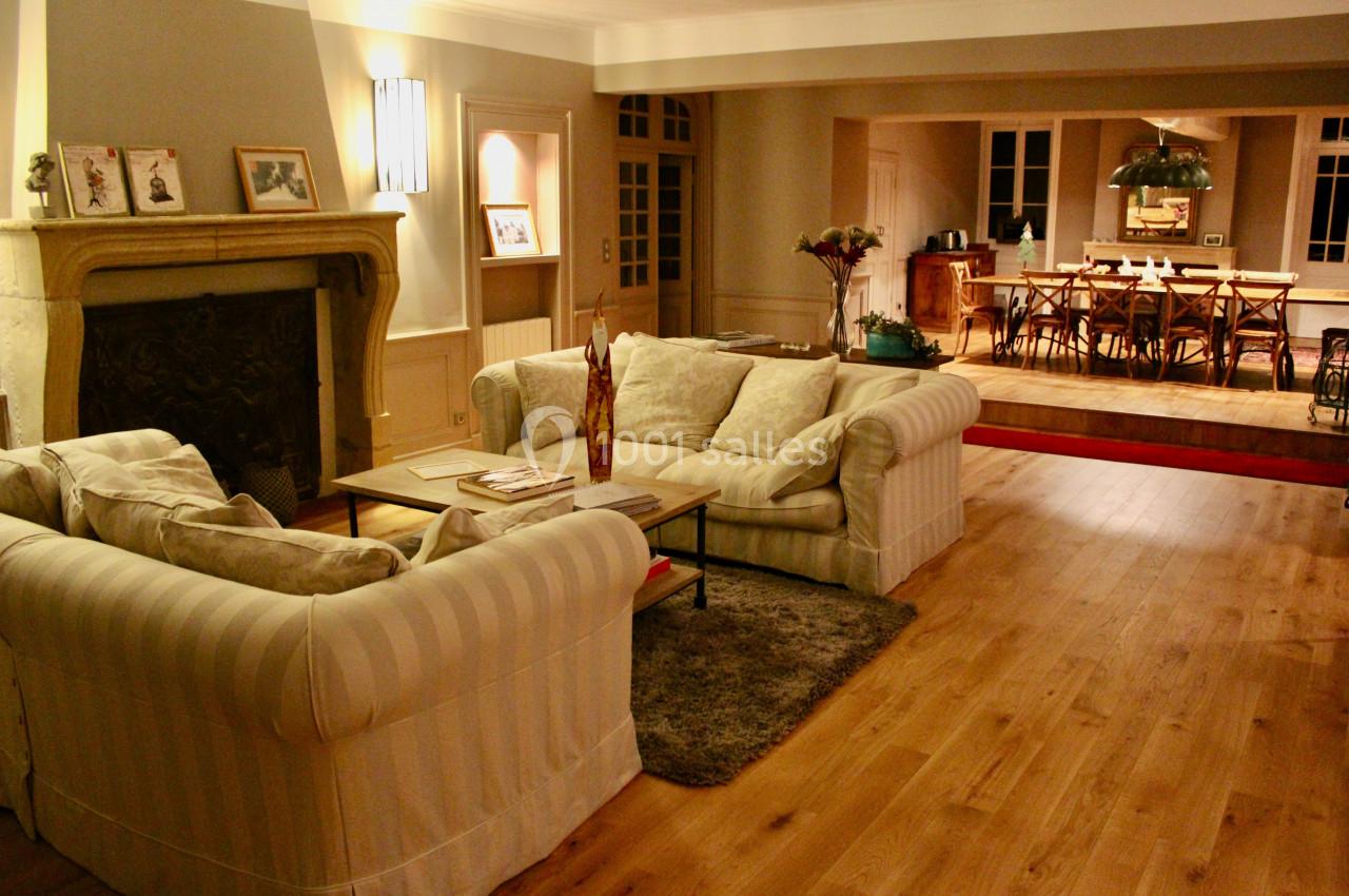 Salon chaleureux avec canapés beiges, cheminée, table basse et salle à manger en arrière-plan sur parquet en bois.