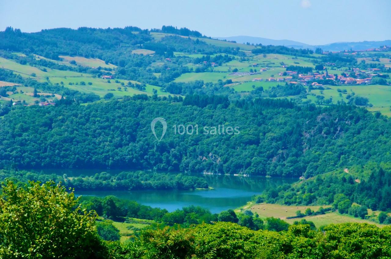 Paysage vallonné avec un lac entouré de forêts et de prairies, village visible au loin sous un ciel dégagé.