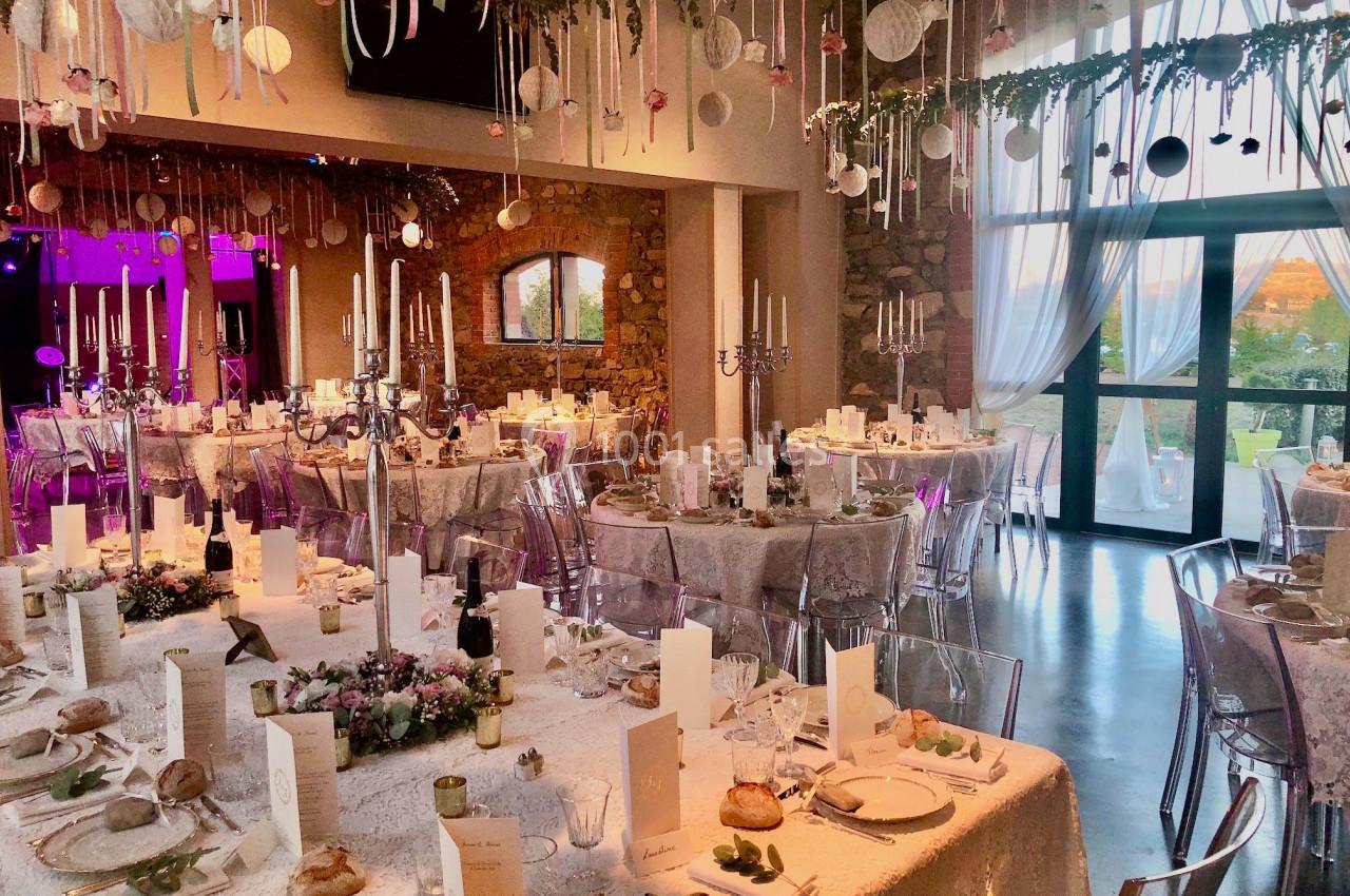 Salle de réception décorée pour un mariage, avec tables dressées, chandeliers et suspensions florales au plafond.