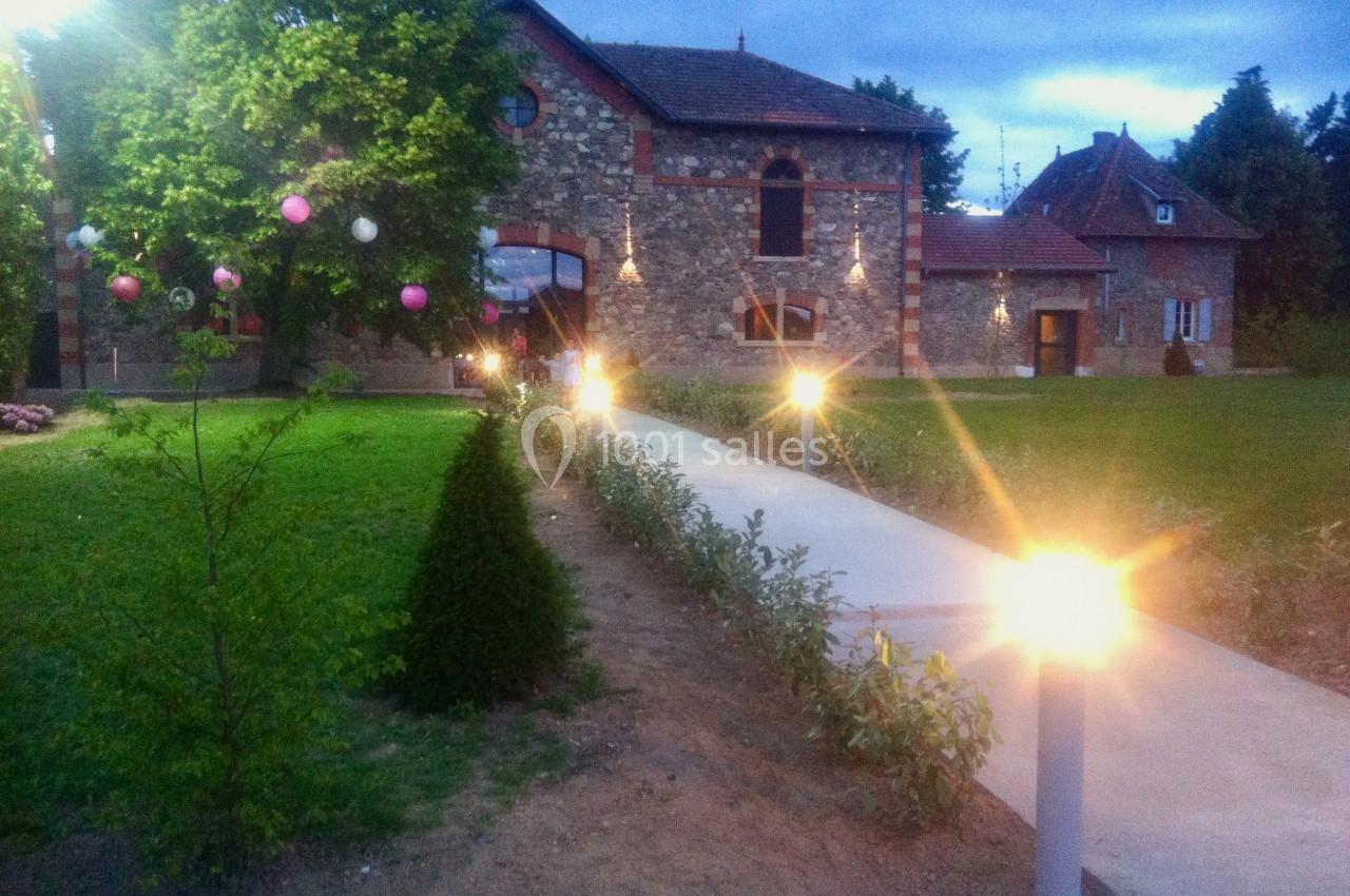 Allée éclairée menant à un bâtiment en pierre entouré de verdure, avec des décorations lumineuses suspendues.