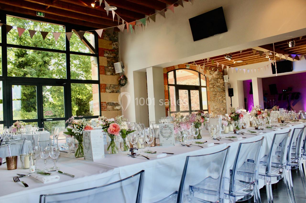 Salle de réception lumineuse avec tables décorées de fleurs et vaisselle, grandes fenêtres donnant sur un extérieur…