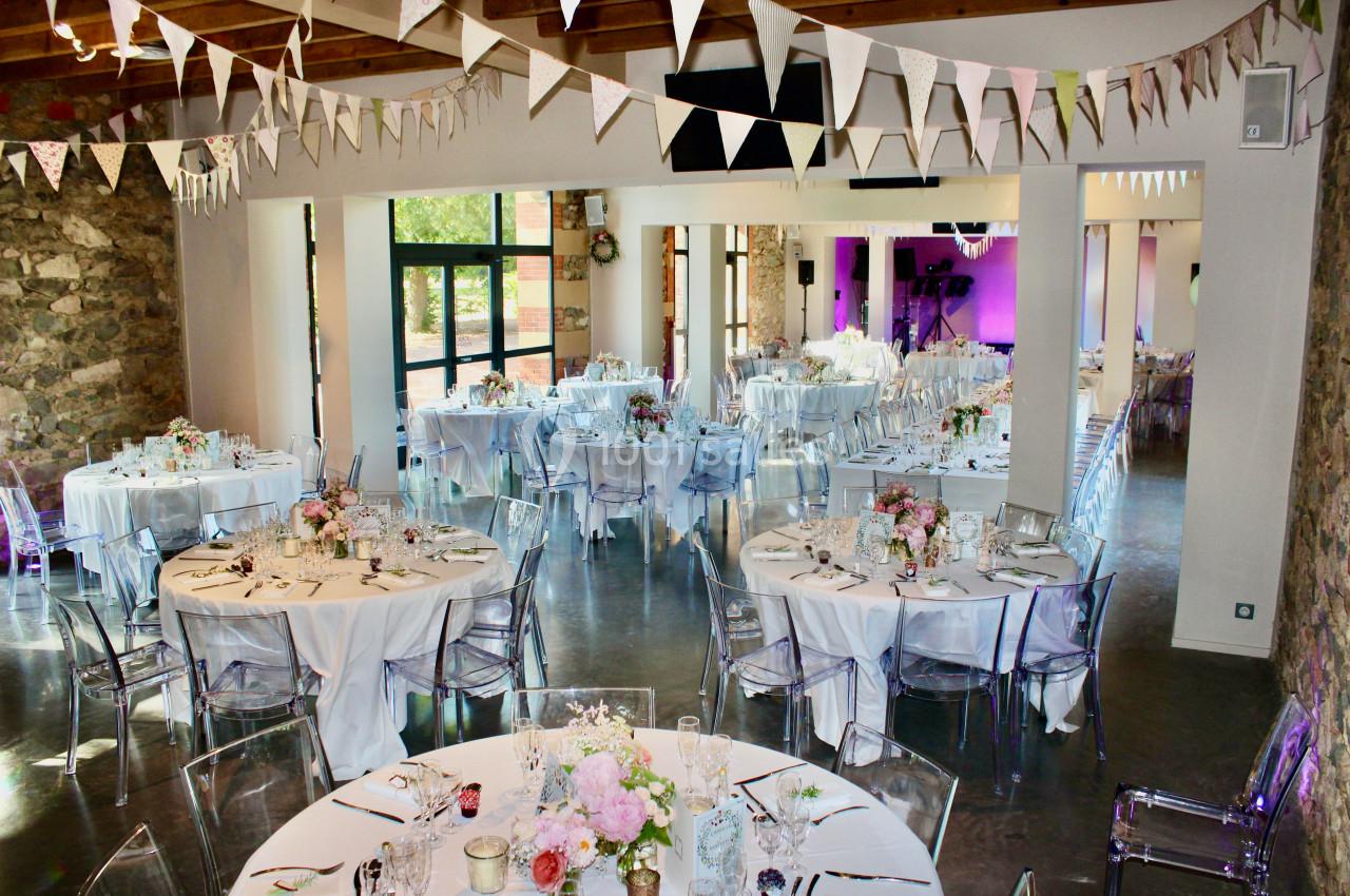 Salle de réception décorée pour un mariage, avec tables rondes dressées, chaises transparentes et guirlandes suspendues.