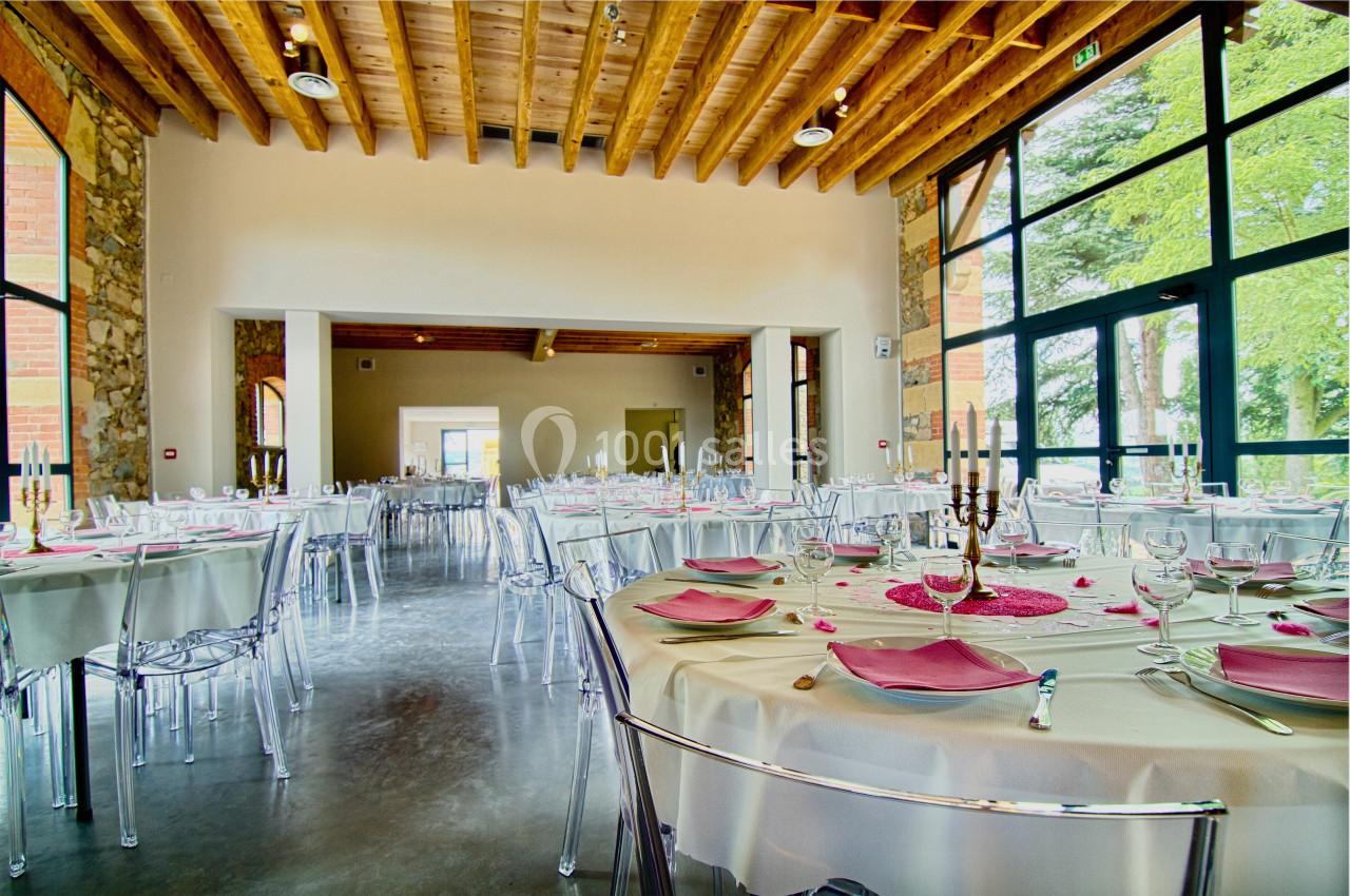 Salle de réception lumineuse avec tables rondes dressées, nappes blanches, serviettes roses et grandes baies vitrées.