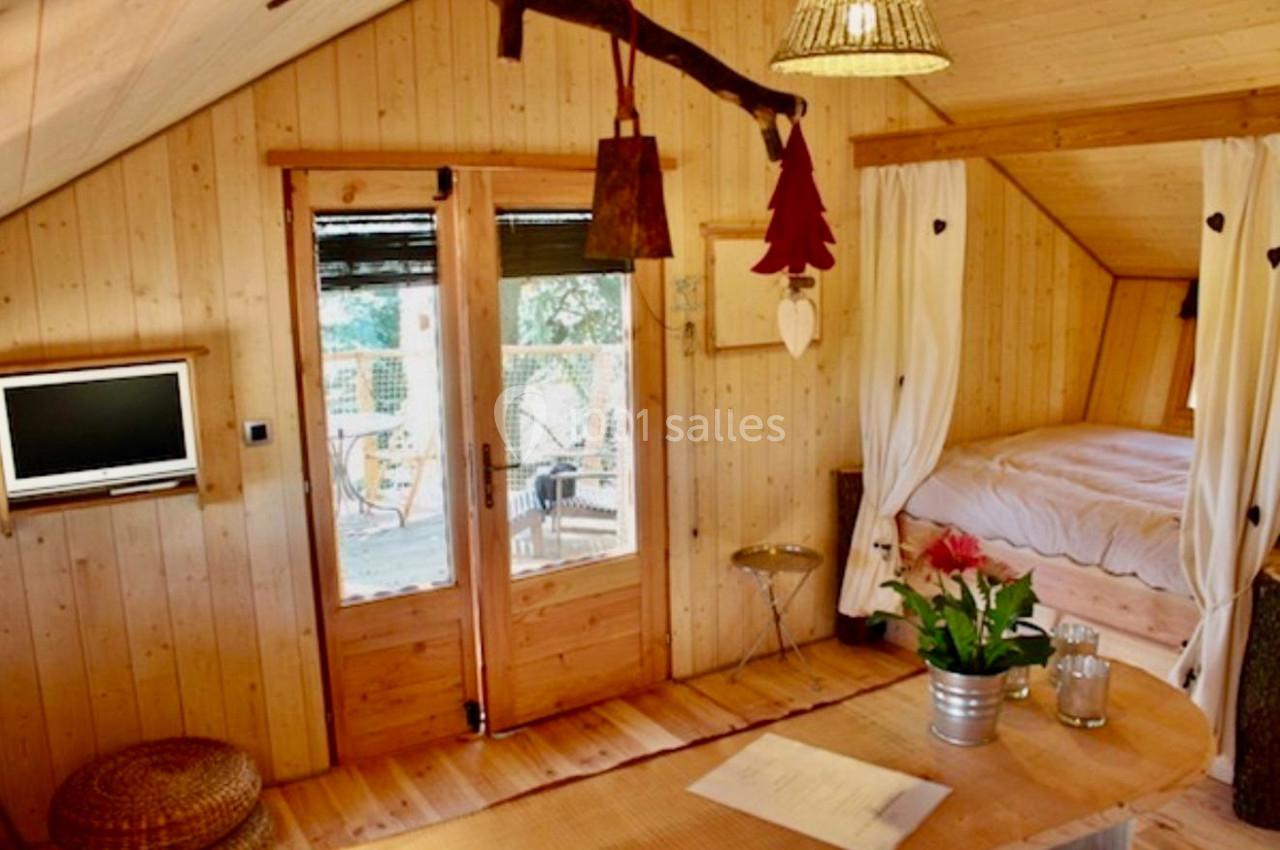 Intérieur d'une cabane en bois avec lit, table, télévision et portes vitrées donnant sur une terrasse.