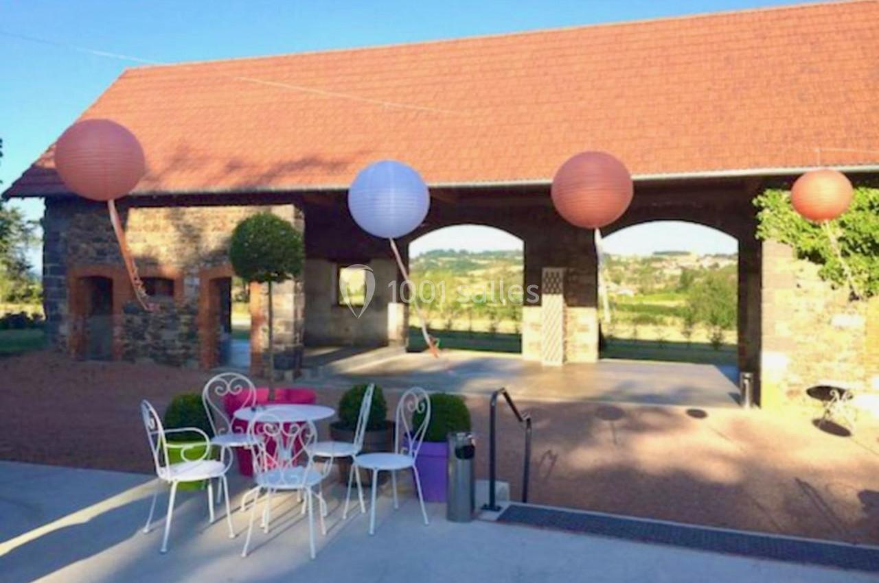 Cour extérieure avec tables et chaises en métal blanc, lanternes suspendues et bâtiment en pierre avec toit en tuiles.