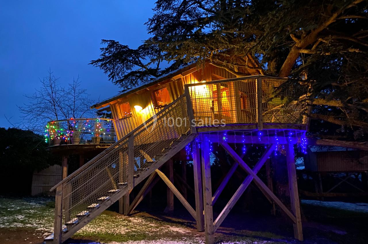 Cabane en bois sur pilotis éclairée, décorée de guirlandes lumineuses colorées, entourée d'arbres en soirée.