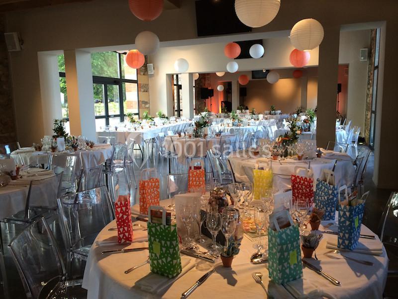 Salle de réception décorée pour un événement, avec tables dressées, lanternes suspendues et lumière naturelle.