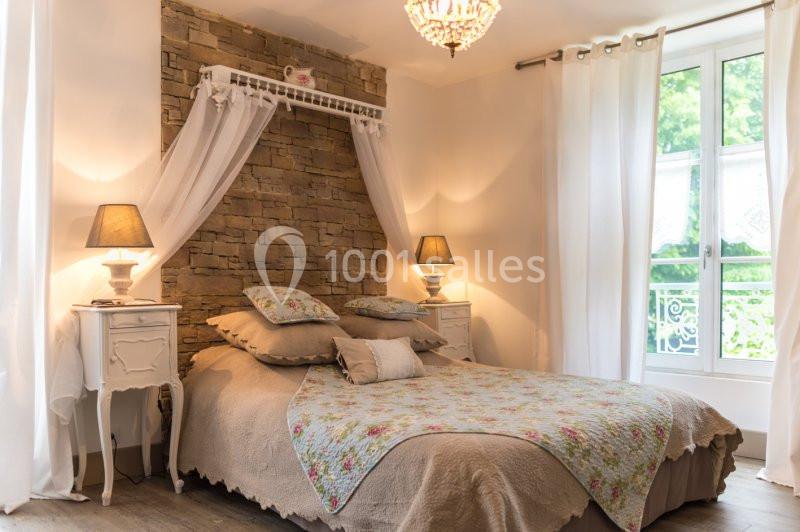 Chambre lumineuse avec lit double, murs en pierre, couvre-lit fleuri, tables de chevet et grandes fenêtres donnant sur un…
