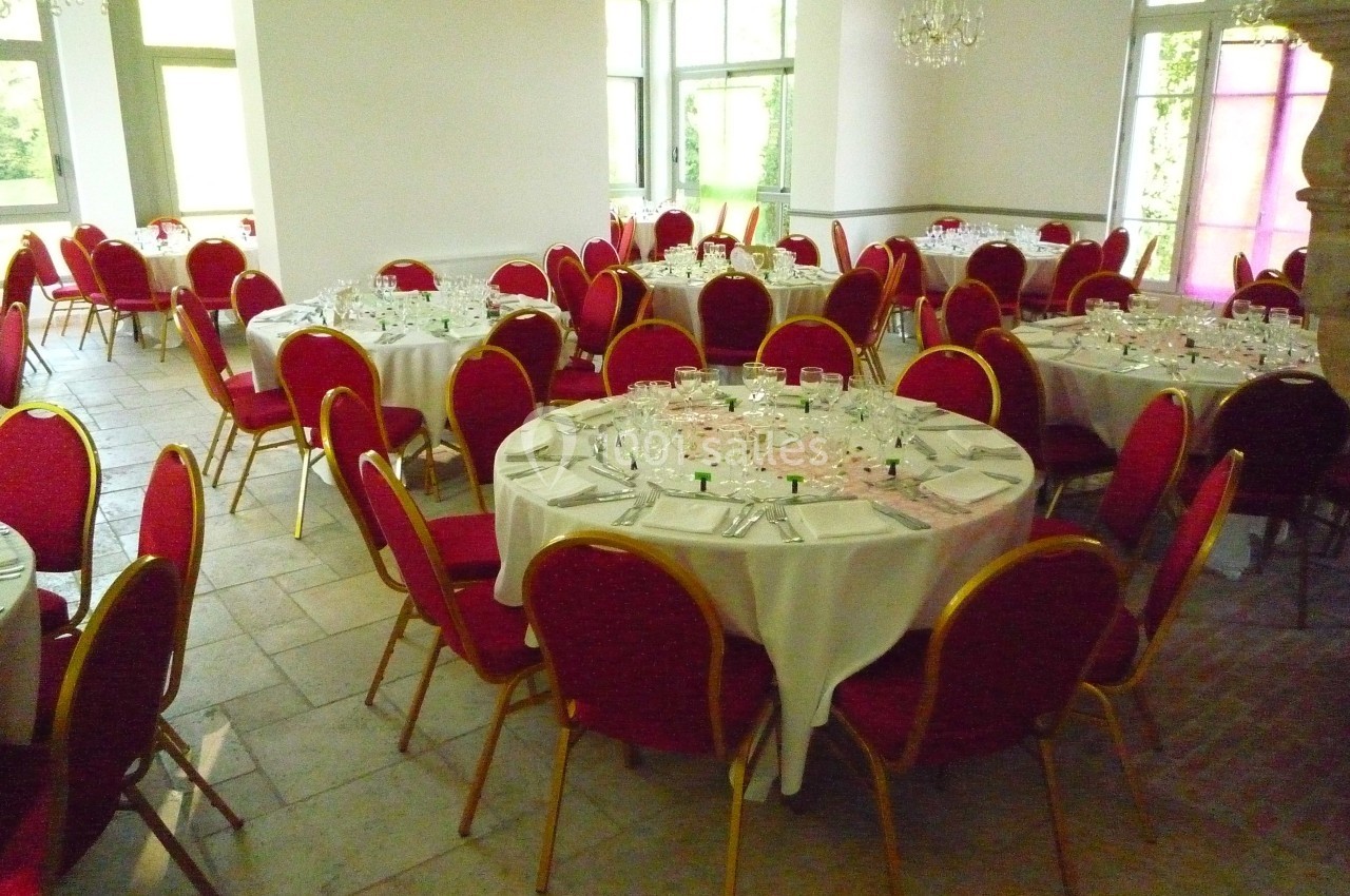 Salle de réception avec des tables rondes dressées, nappes blanches et chaises rouges, éclairée par la lumière naturelle.