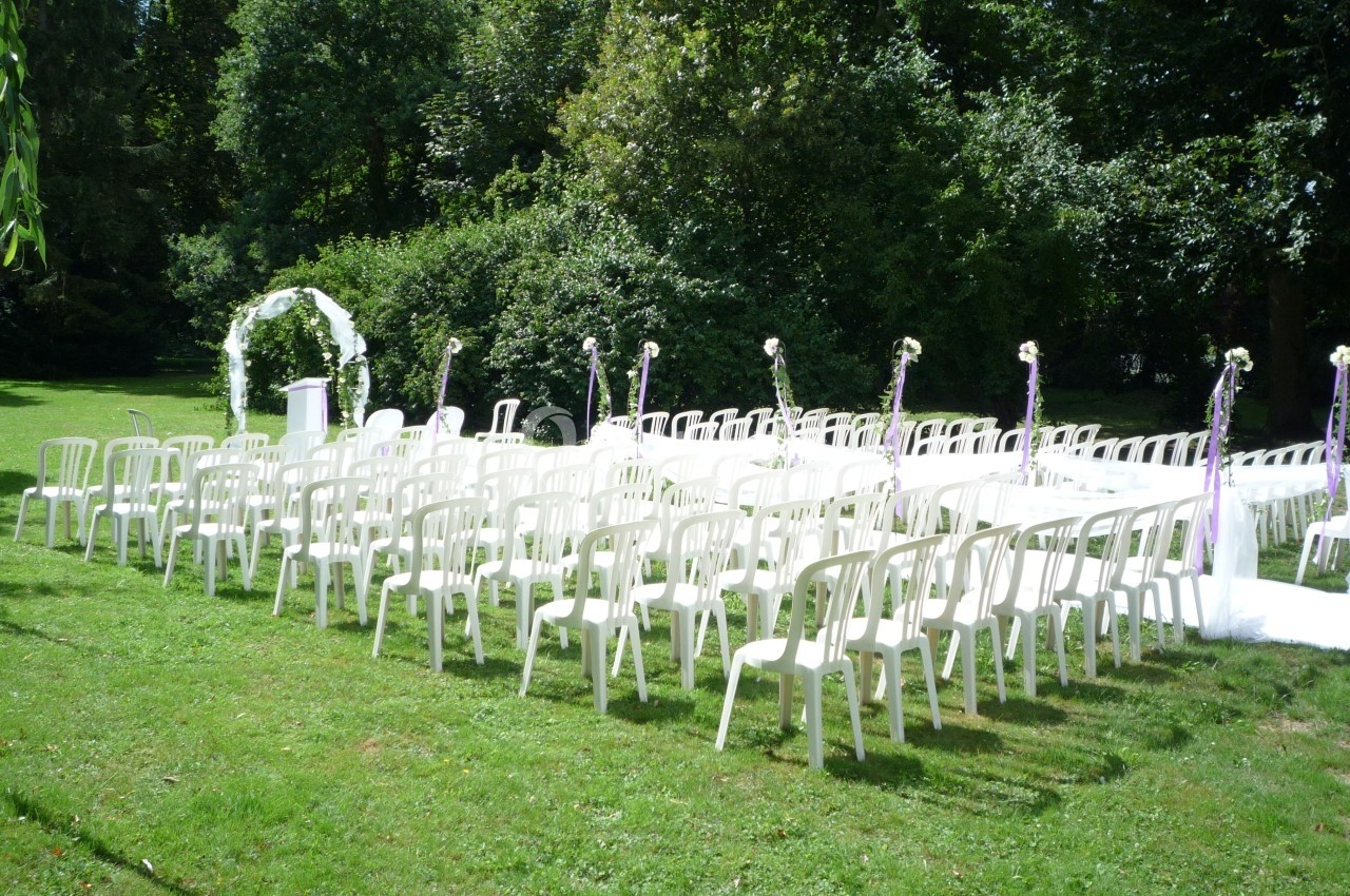 Chaises blanches disposées en rangées sur une pelouse, face à une arche décorée pour une cérémonie en plein air.