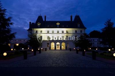 Vue aérienne d'un bâtiment en forme de cour rectangulaire avec des toits en tuiles, entouré de champs et d'arbres.