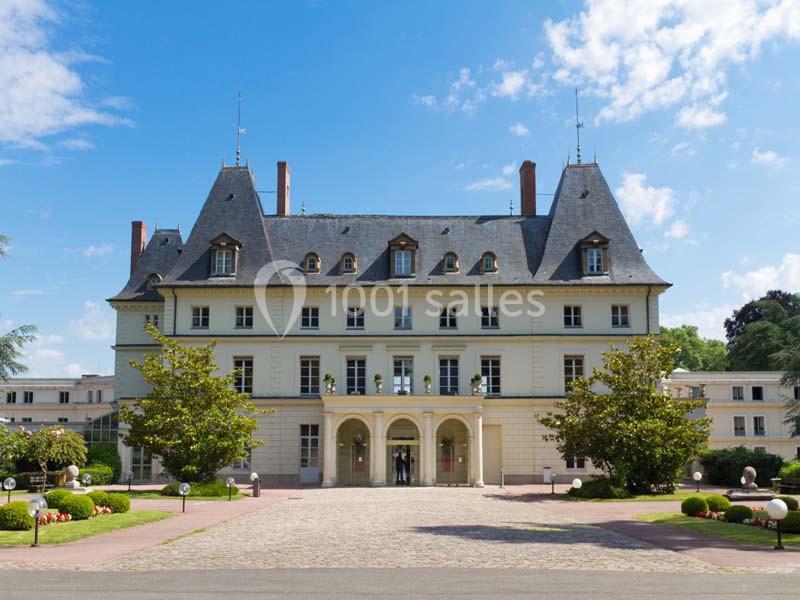 Façade d'un grand bâtiment de style classique avec toit en ardoise, entouré de jardins et d'arbres sous un ciel bleu.