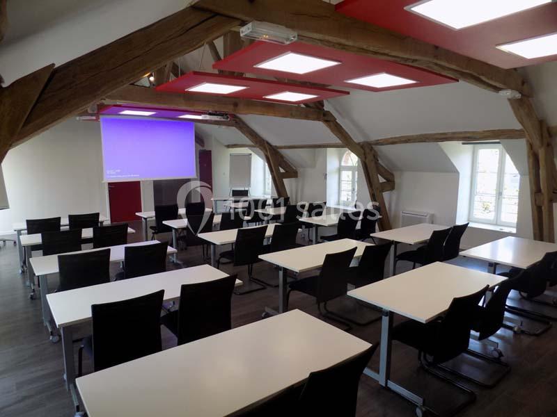 Salle de formation lumineuse avec poutres apparentes, tables blanches, chaises noires et écran de projection allumé.