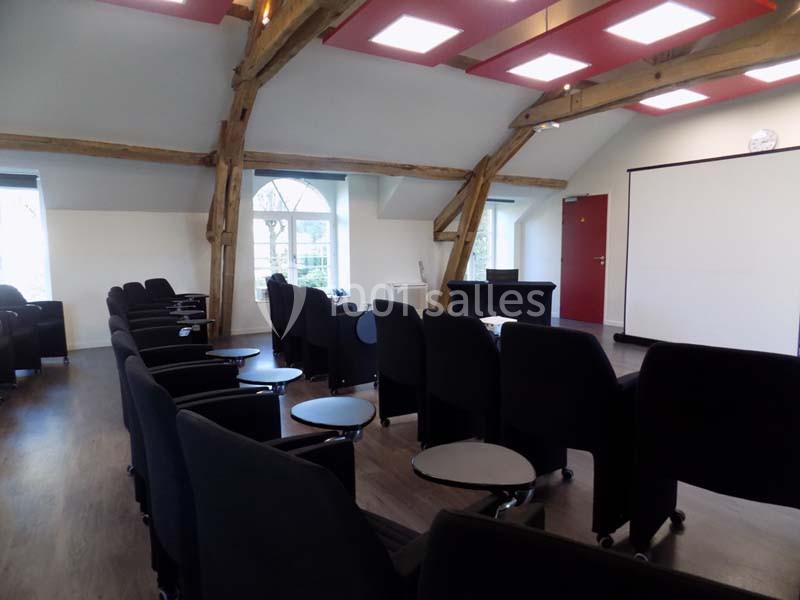 Salle de réunion lumineuse avec fauteuils noirs, tables individuelles, poutres apparentes et écran de projection.