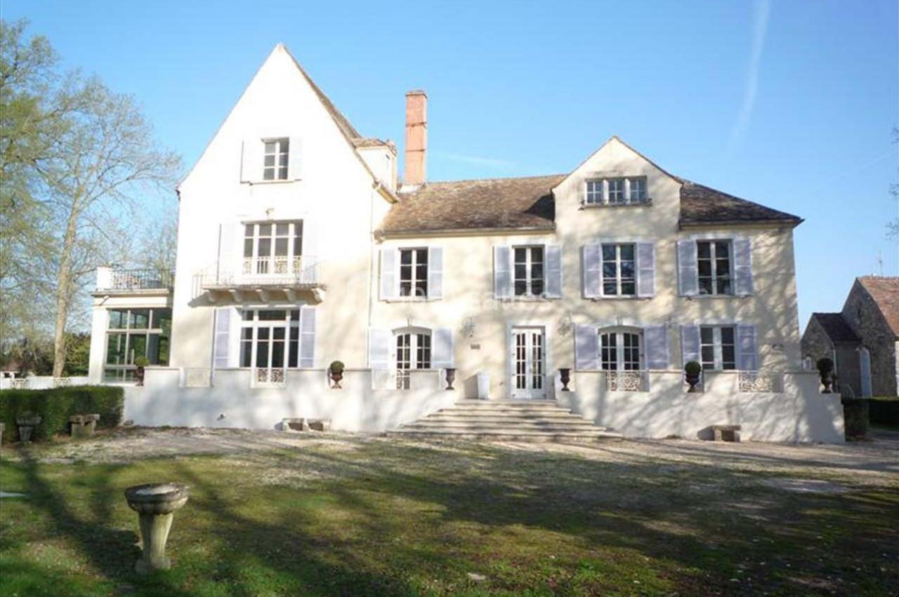 Façade d'une grande maison de campagne en pierre claire avec jardin et allée devant.