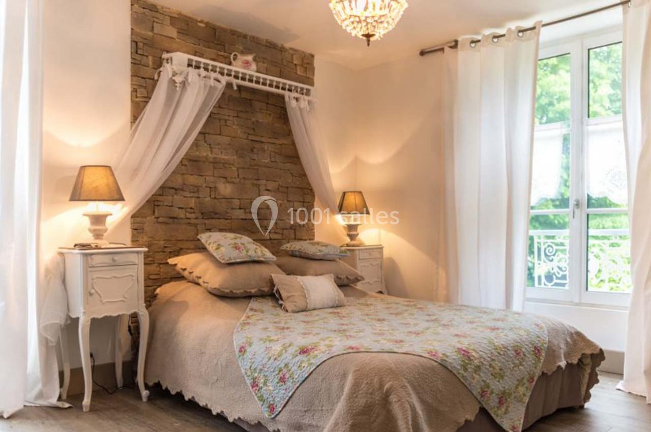 Chambre lumineuse avec lit double, tête de lit en pierre, luminaires et grande fenêtre donnant sur un jardin verdoyant.