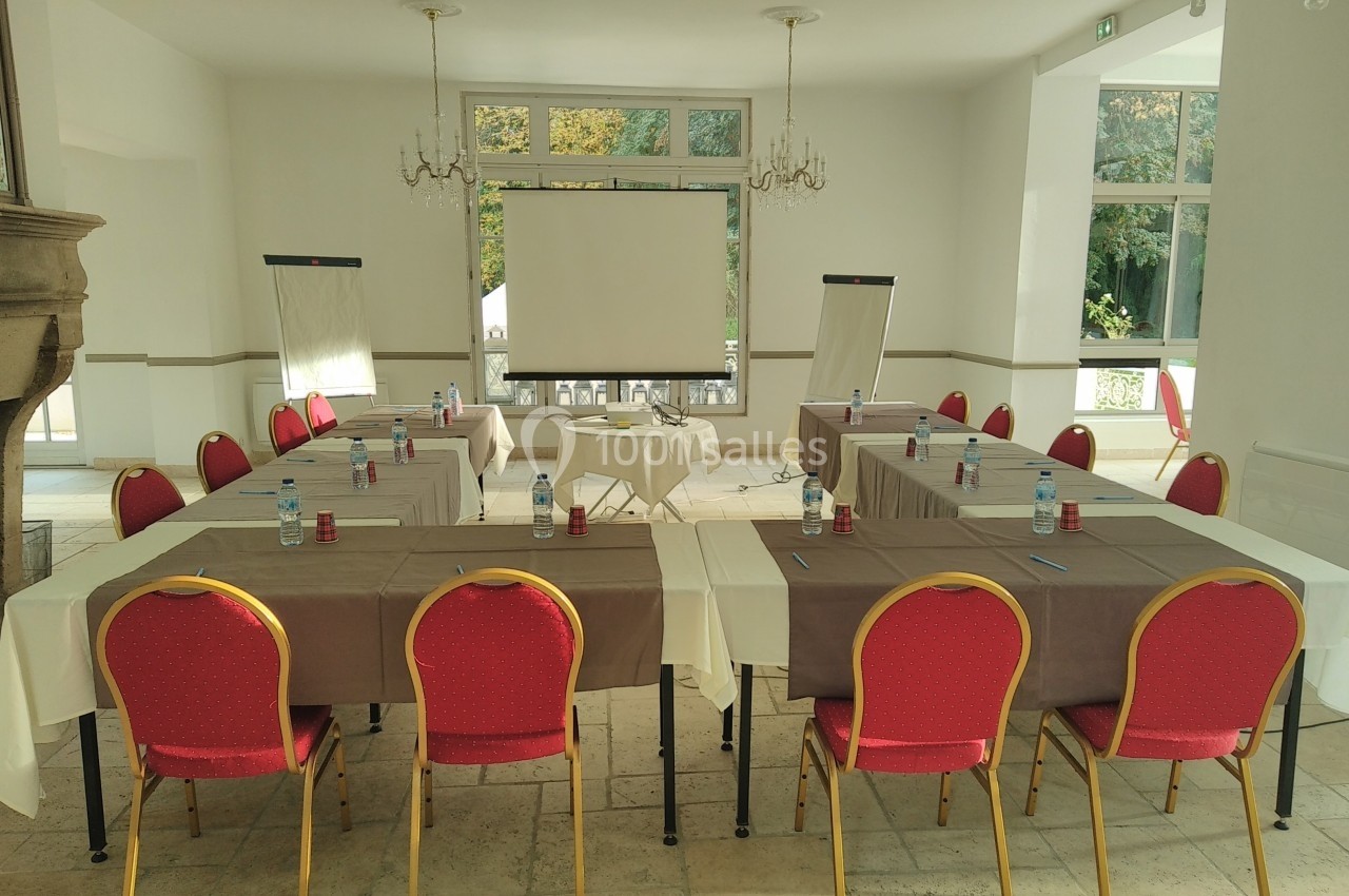Salle de réunion lumineuse avec tables disposées en U, chaises rouges, paperboards et écran de projection.