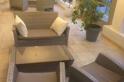 Mobilier de jardin en rotin avec coussins, disposé dans une véranda lumineuse avec des plantes en pot.