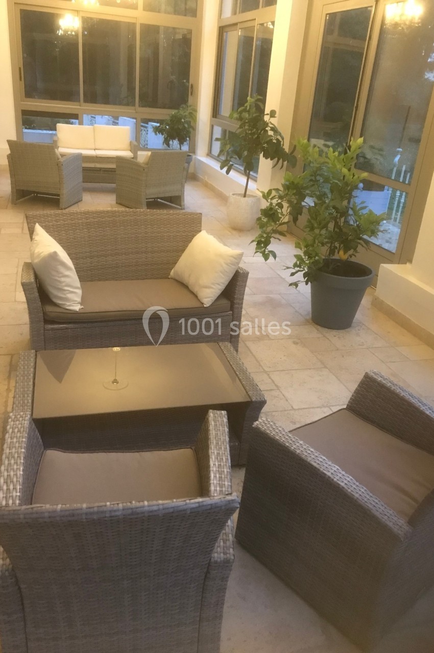 Mobilier de jardin en rotin avec coussins, disposé dans une véranda lumineuse avec des plantes en pot.